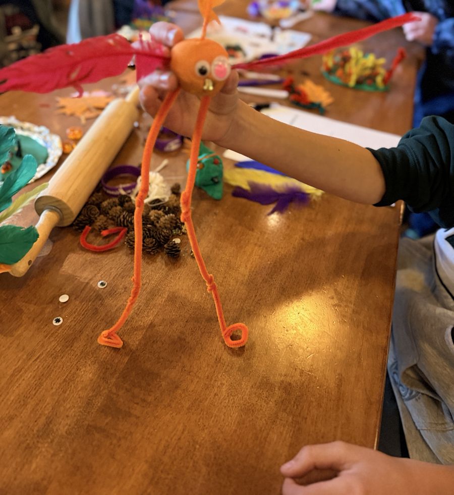 A child holds up a playdough bird with pink feather wings and pipe cleaner legs